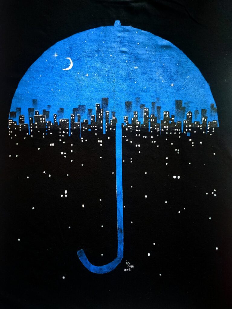 Hand-painted T-shirt design featuring a stylized blue umbrella forming a dome over a city skyline at night. The umbrella's handle extends down like the letter 'J,' blending seamlessly into the black background. Inside the dome, a crescent moon and stars shine above glowing blue buildings, creating a dreamy, protective atmosphere. The artwork is signed 'in me art' at the bottom