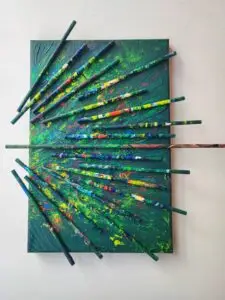 A mixed media abstract artwork featuring a small green-painted canvas with a dynamic array of colorful sticks arranged in a radial explosion. The sticks, each uniquely covered in vibrant layers of yellow, blue, red, and green paint, protrude from the surface at various angles. The textured background echoes the same vivid hues, creating a sense of energetic movement and depth. The composition evokes a sense of chaos turned into harmony, as if capturing a colorful explosion frozen in time.